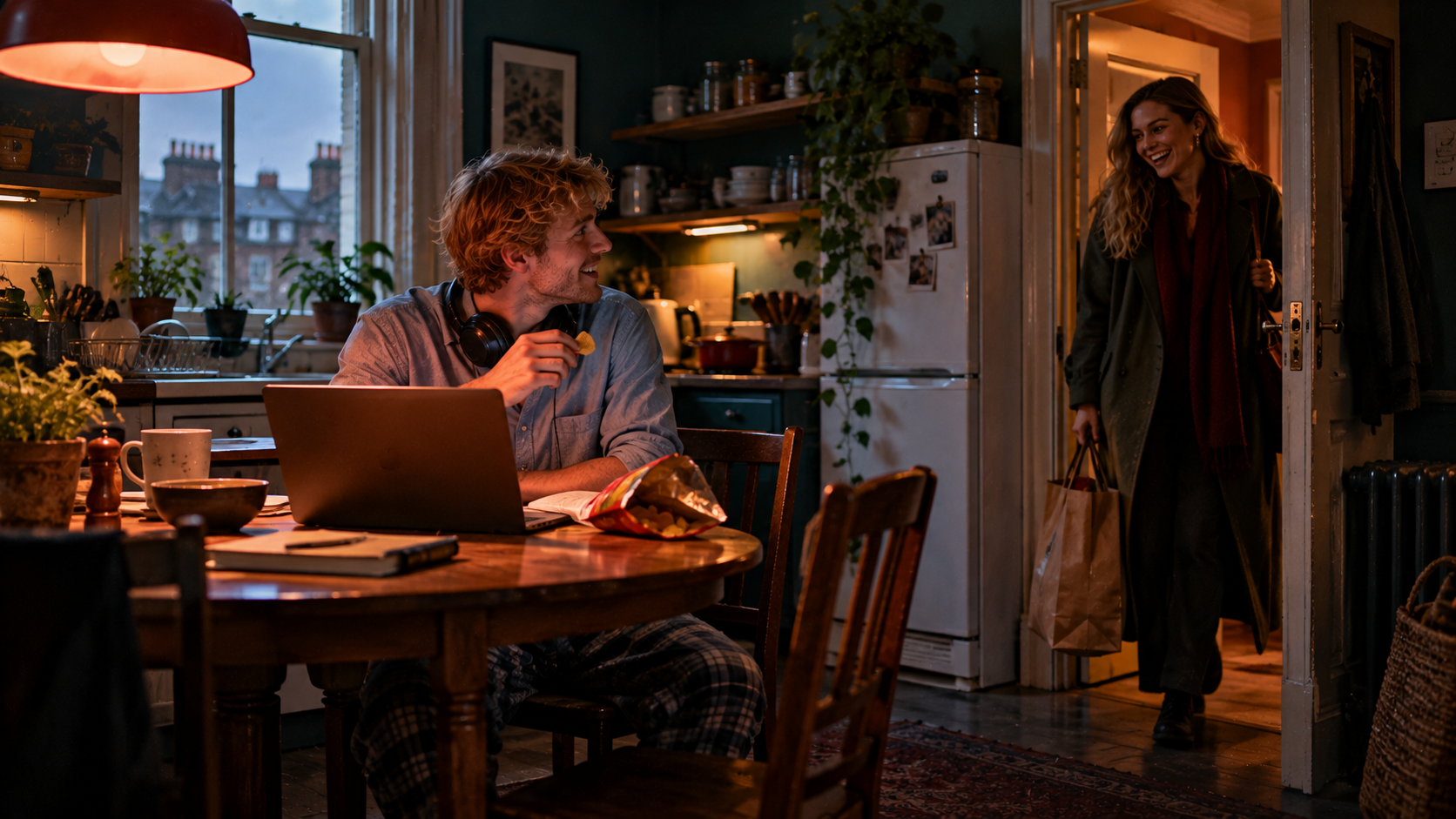 Afternoon in a small shared flat: a young man in a smart shirt and pyjama trousers sits at the kitchen table in front of a laptop with headphones round his neck, a packet of crisps next to him, while his flatmate, a young woman in a coat with a shopping bag, stops in the doorway and laughs; grey daylight from the window, warm light over the table